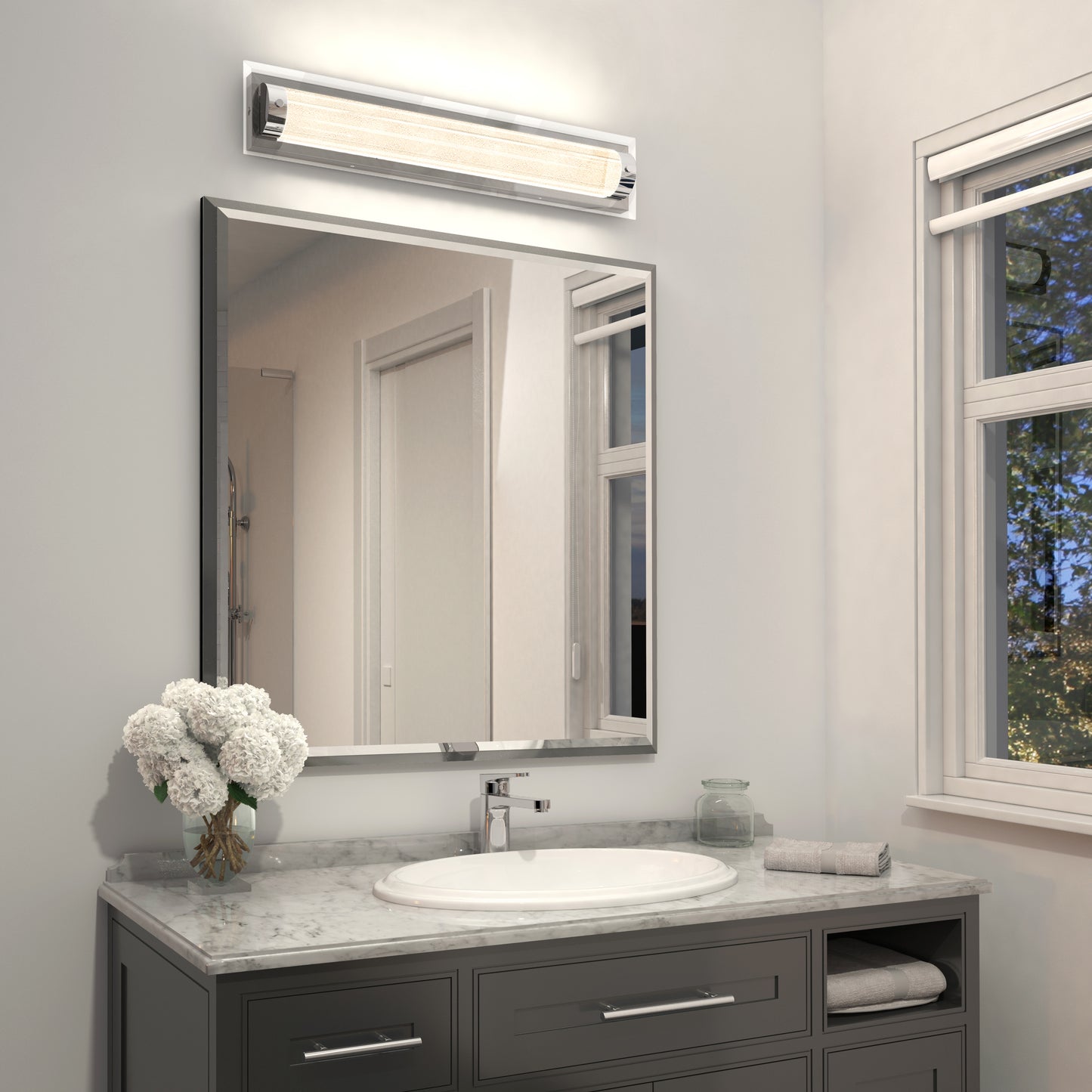 A modern bathroom featuring a marble countertop, oval sink, rectangular mirror, and a vase of white flowers. Above the mirror is the Artika Glamour LED bathroom vanity light 3 CCT. Natural light enters from a window on the right.