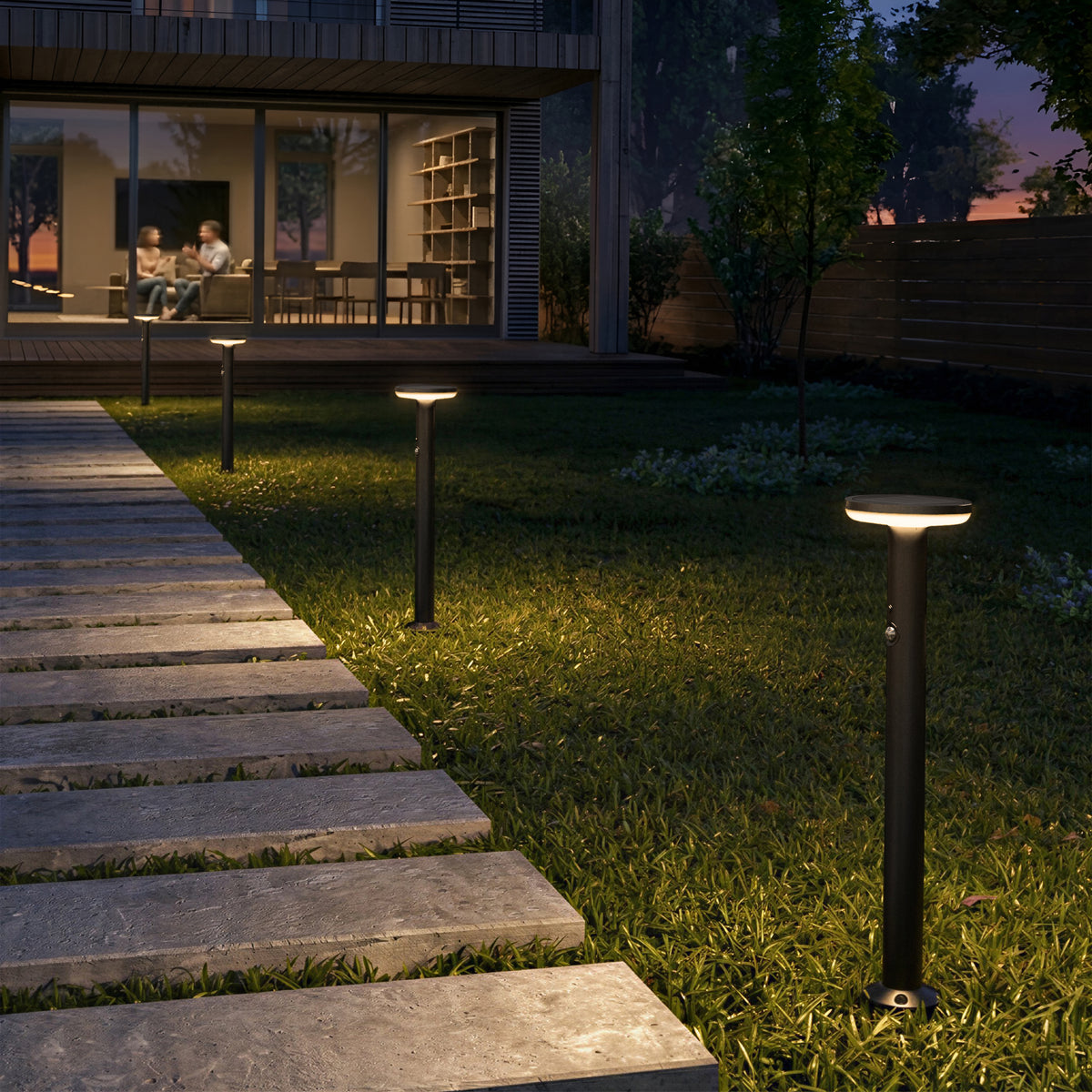 A modern backyard at dusk lit by Artika Urbia Solar LED Bollards, featuring grass, trees, and two people on a patio inside.