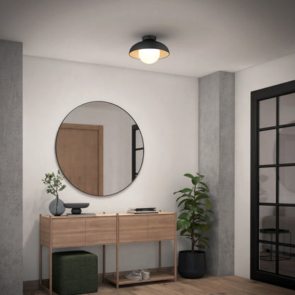 A modern entryway with a wooden console table, round mirror, potted plant, green ottoman, and black-framed glass door is illuminated by the Artika Boden LED flush mount light 5 CCT. Minimalist décor completes the look.