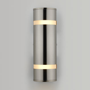 The Artika Q1 indoor-outdoor wall light features a modern cylindrical design with a brushed metal finish and two horizontal bands, emitting warm white light at the top and bottom, mounted on a plain light gray wall.