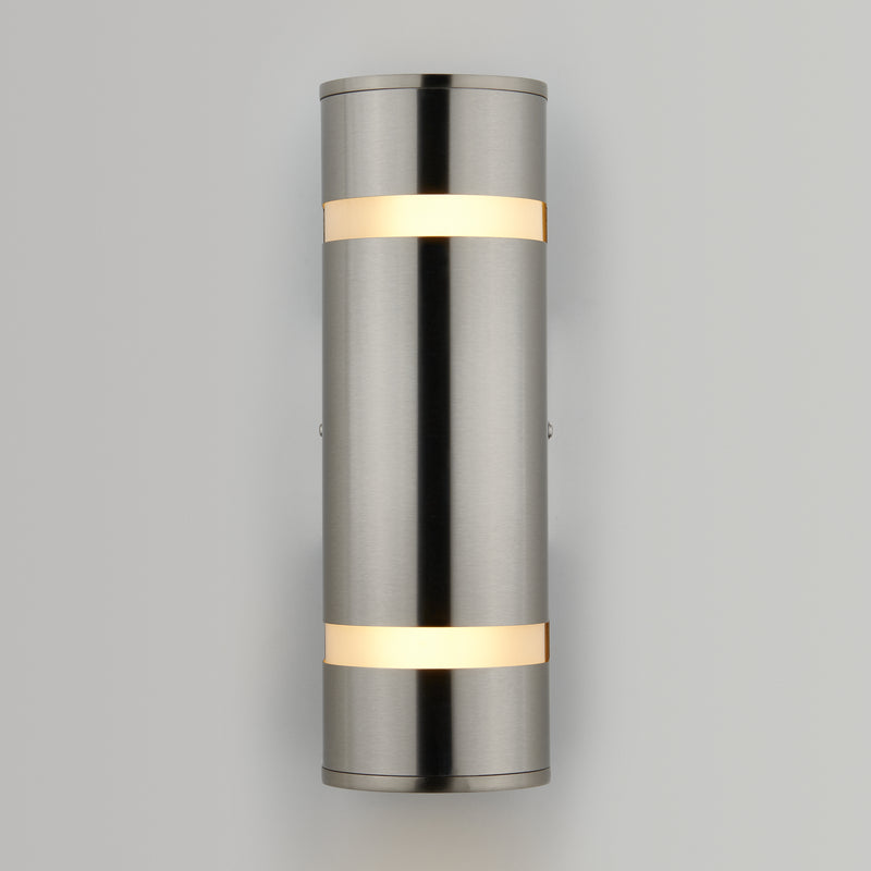 The Artika Q1 indoor-outdoor wall light features a modern cylindrical design with a brushed metal finish and two horizontal bands, emitting warm white light at the top and bottom, mounted on a plain light gray wall.