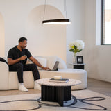 A man sits on a cream sofa in a modern living room, holding a mug and looking at a laptop. A round coffee table and a side table with flowers are in front of him. The room is bright and minimalistic.