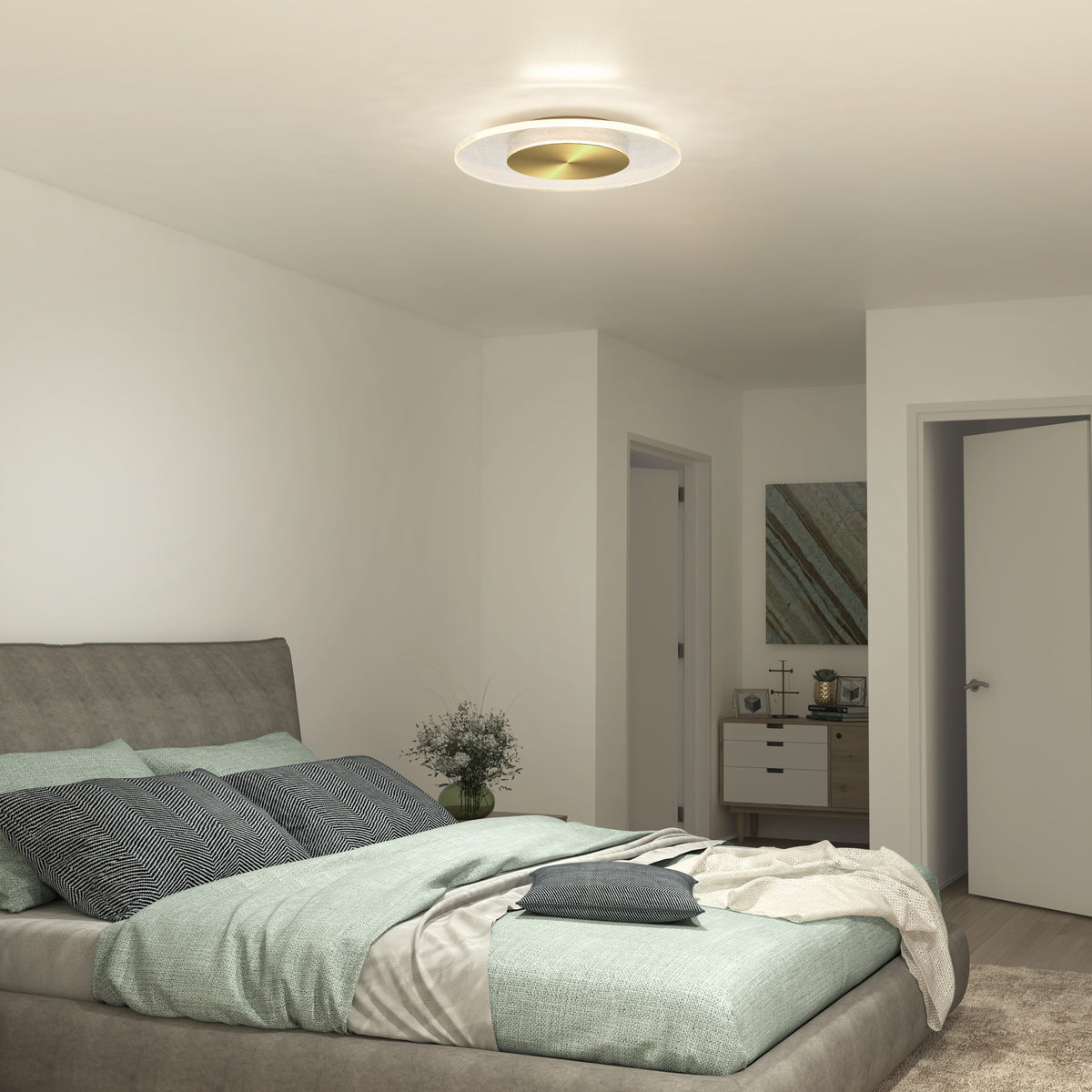 A modern bedroom with a gray upholstered bed, green and gray bedding, a nightstand with flowers, Artikas Essence Disk LED flush mount ceiling light, white walls, two doors, and a dresser with decor.