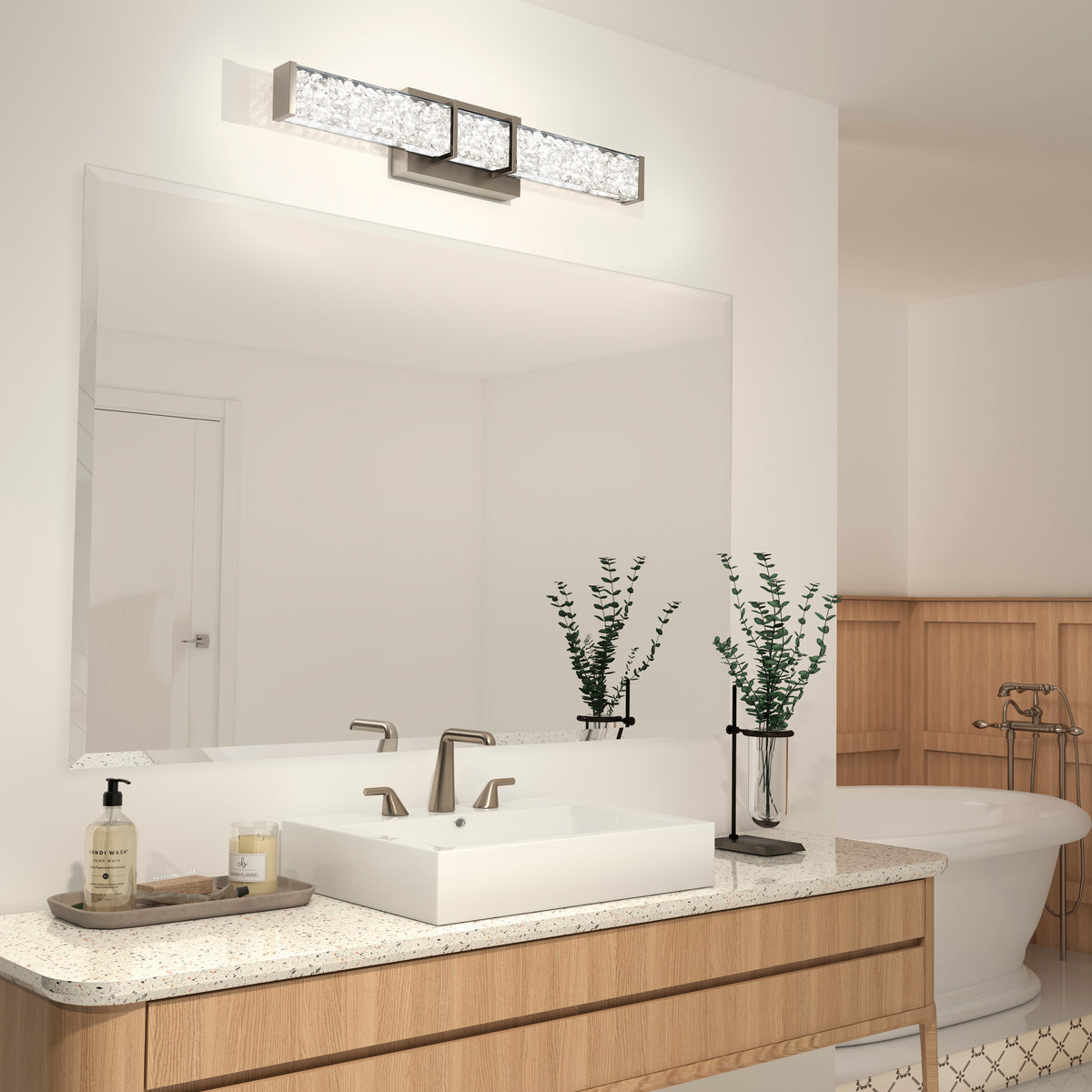 Modern bathroom with a large mirror above a light wood vanity, a rectangular sink, and elegant faucet. Two glass vases with leafy green stems and a soap dispenser are on the countertop. A bathtub is visible in the background.