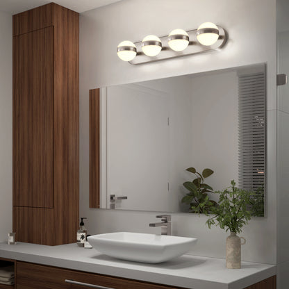 Modern bathroom with a large mirror, vessel sink, wooden cabinets, a plant in a vase, soap dispenser, and a four-globe light fixture above the mirror. A window with blinds is on the right.
