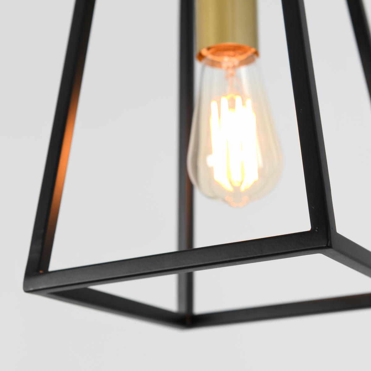 A close-up of a glowing vintage-style light bulb in a modern, geometric black metal pendant lamp with brass accents, set against a plain white background.
