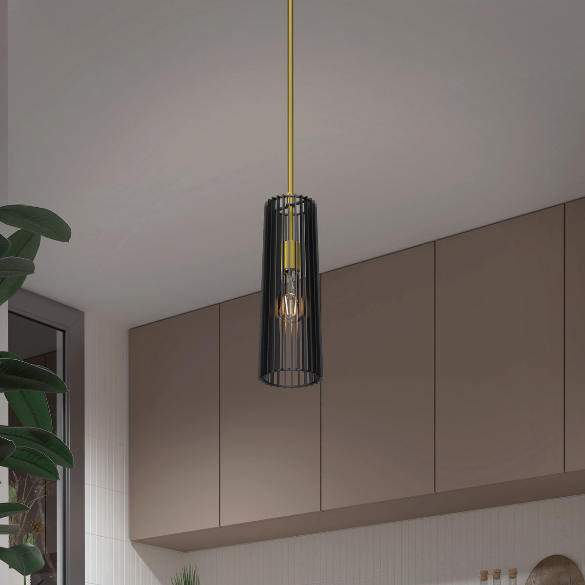 The Artika Gatsby Mini modern mid-century pendant light, featuring a black cylindrical cage shade and gold fixture, hangs in a contemporary kitchen with beige cabinets and a large green plant nearby.