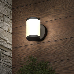 The Artika Landon LED Outdoor Wall Light 2-Pack brightens wood siding near stone, casting light on a leafy green plant below.