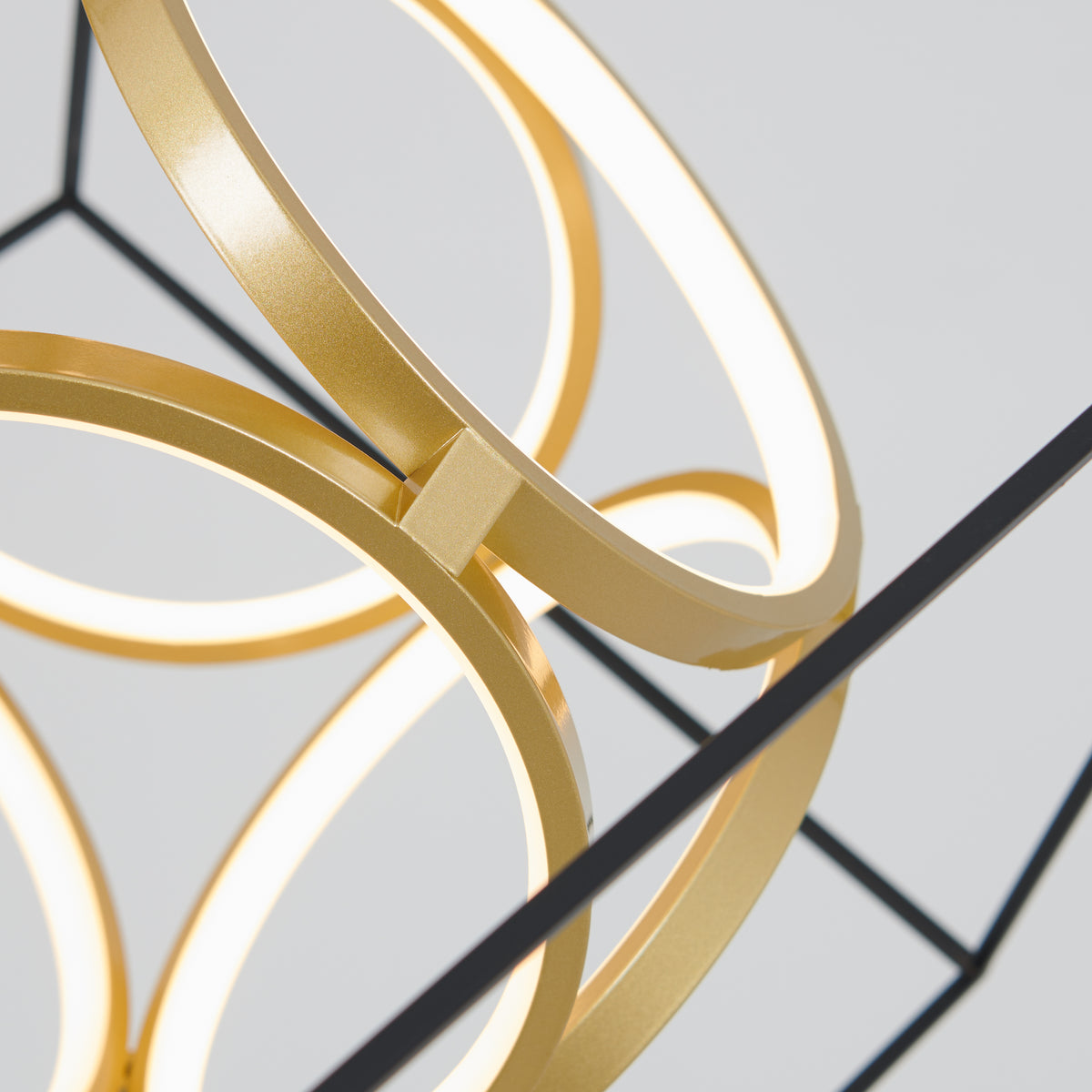 Close-up of a modern light fixture featuring intersecting golden rings with integrated LED lights, set against a plain light gray background. The design appears geometric and contemporary.