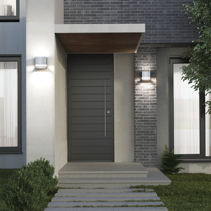 Modern house entrance featuring the Valor Square LED indoor/outdoor wall light by Artika-CAN-dev, a dark front door, brick and concrete walls, a large curtained window, and a small garden with shrubs and grass along a stone path.