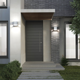 Modern home entryway with a dark gray door, brick and concrete walls, small porch roof, window with white curtains, landscaped greenery, and two Artika Valor Square LED indoor-outdoor wall lights.