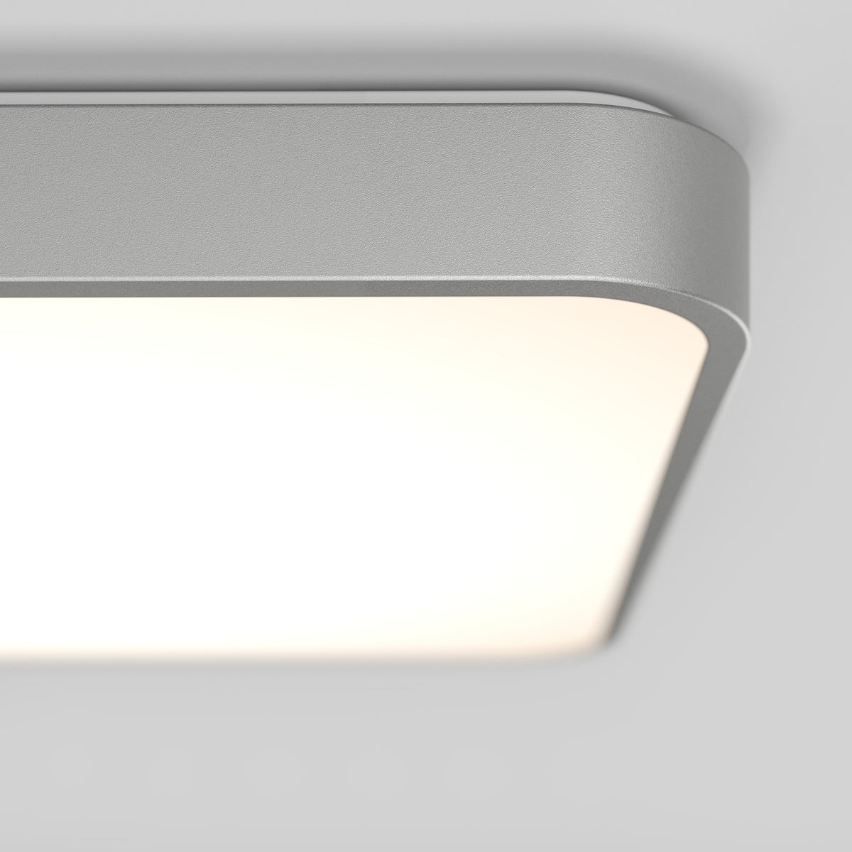 Close-up of a modern, rectangular ceiling light fixture with rounded edges, emitting a soft white glow. The fixture has a smooth, metallic frame and is mounted on a light gray ceiling.