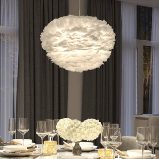 A modern dining table set with glassware and plates sits beneath the Artika Plume LED pendant light. The scene is accented by two white flower arrangements, dark curtains, and large windows.