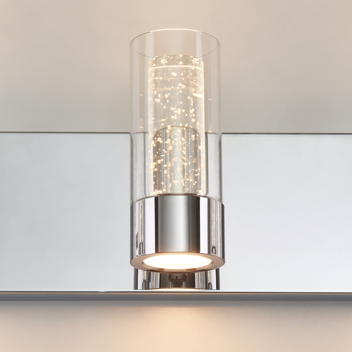 A modern wall sconce with a cylindrical glass cover, chrome base, and small LED lights inside, mounted on a mirrored surface.