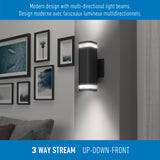 A modern black wall light fixture emits light upward, downward, and forward, illuminating part of a living room wall decorated with framed art above a gray couch. Text describes its multi-directional design.