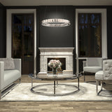A modern living room with a marble fireplace, round glass coffee table, white sofas, and a circular chandelier. A vase of pink flowers and books sit on the table, with large windows on either side of the fireplace.