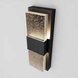 A modern wall sconce with a black rectangular base and two frosted glass panels featuring a scattered dot pattern, emitting a soft, warm light.