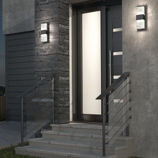 Modern house entrance with gray stone and concrete walls, black metal railings, and Artika Mazza Frosted indoor-outdoor LED wall lights flanking a frosted glass front door. Steps lead up to the entrance.