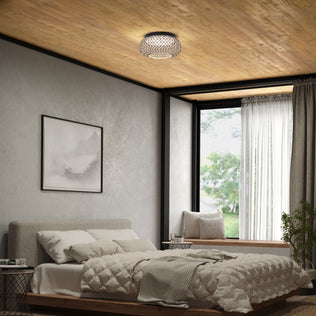 A cozy bedroom features a large bed with neutral bedding, a framed picture, a window seat with cushions, and an Artika Benson 15 in. LED flush mount light 5 CCT on the wooden ceiling. Sunlight pours through sheer-curtained windows.