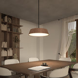 A modern dining room with a wooden table, four upholstered chairs, an open book and cup on the table, a large pendant light above, built-in shelves with books and decor, and a window with sheer curtains.