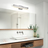 Modern bathroom with a white vessel sink on a marble countertop, wall-mounted faucet, large mirror, and decorative green vases with greenery. A sleek, horizontal light fixture is above the mirror.
