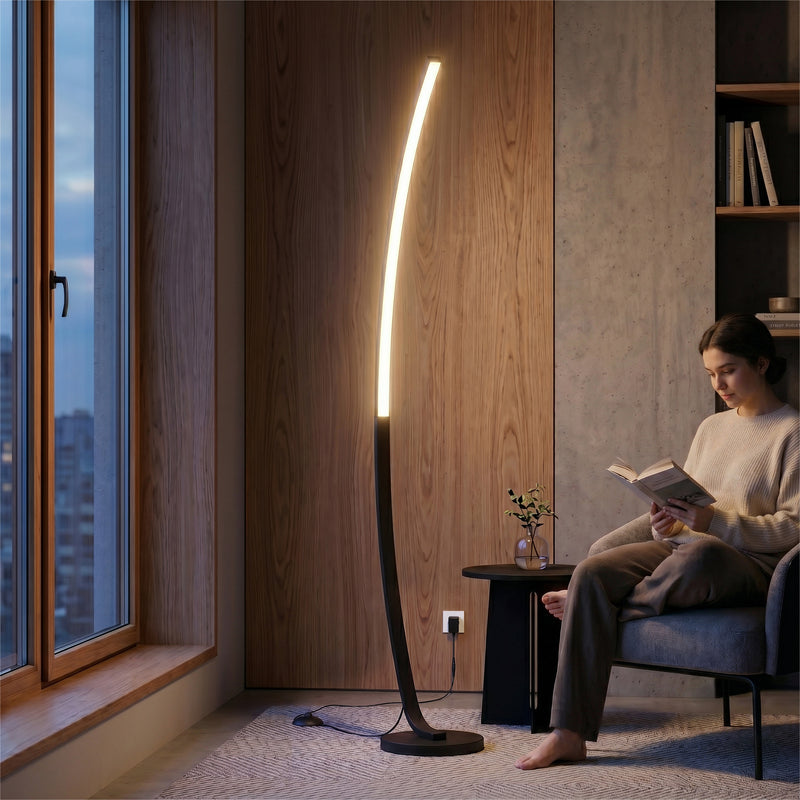 A woman reads by the Artika Arc LED Floor Lamp, featuring an arched design and dimmable lighting. The warm glow illuminates the wood-paneled, city-view living room with its sleek, minimalist decor.