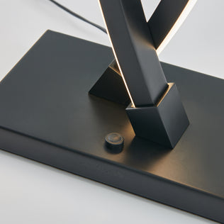 Close-up of the Artika Wave LED Table Lamp’s modern black base with a round button switch. Two angled stems with integrated LED strips intersect above, and the sleek surface features subtle Artika branding.