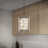 A modern kitchen with light wood cabinets features a hanging pendant light with a cylindrical glass and crystal shade, emitting a bright, elegant glow above a light countertop.