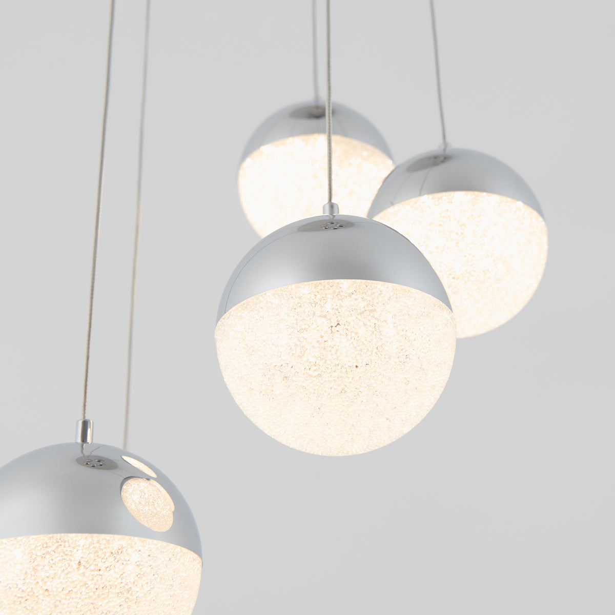 Four modern spherical pendant lights with shiny silver tops and textured, glowing white bottoms hang at different heights against a plain light gray background.
