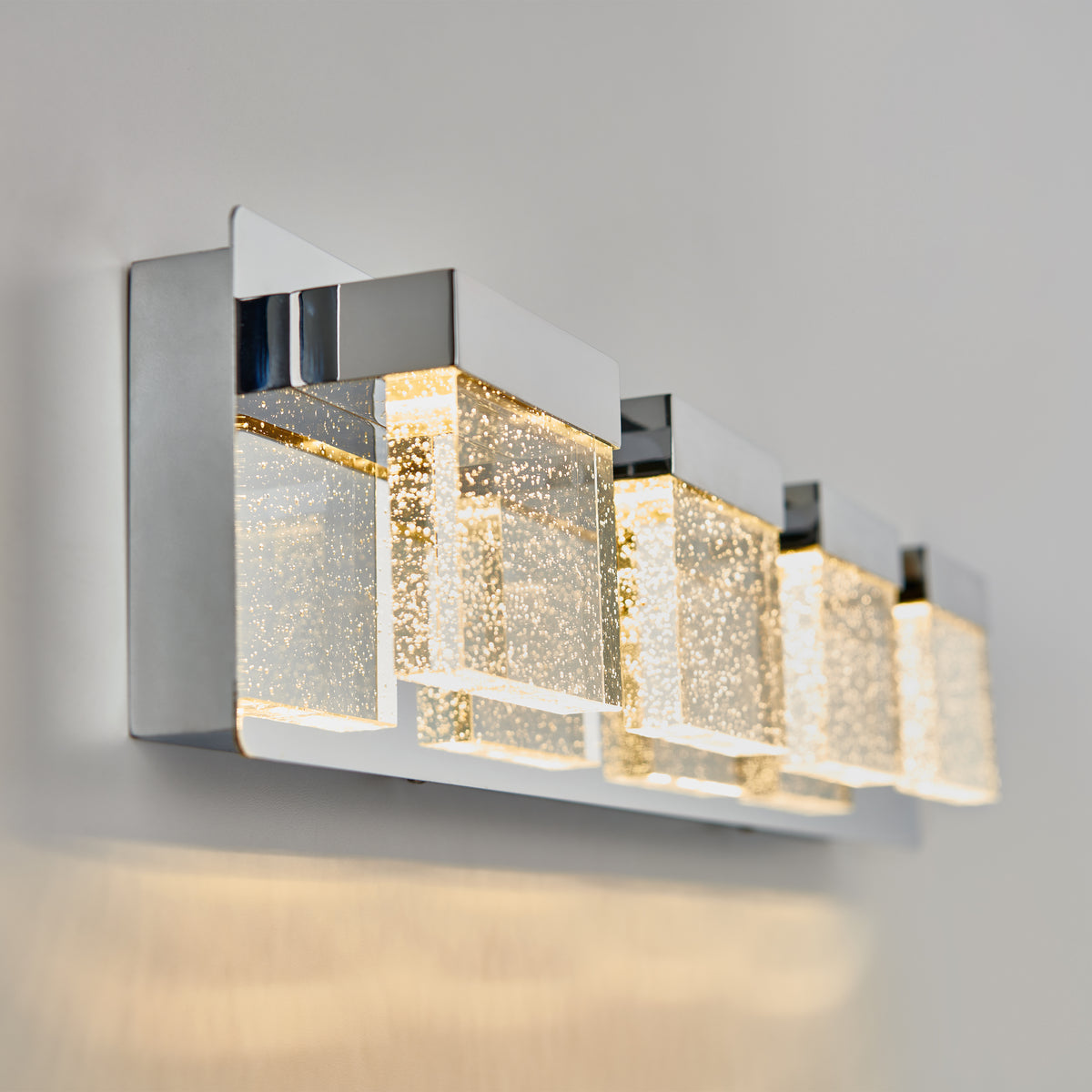 A modern wall sconce with four rectangular LED lights featuring chrome accents and bubble-like textures inside the glass, casting a soft, warm glow on a light-colored wall.