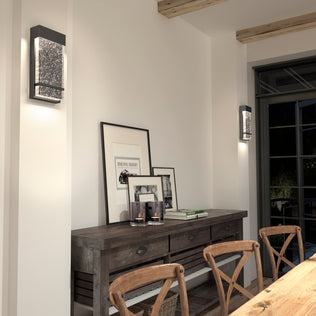 A rustic dining room features wooden chairs and table, framed art leaning on the tabletop, candles above, and Artikas Skyler indoor-outdoor LED wall lights mounted on the walls. Large windows fill the space with natural light.