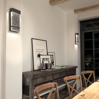 A rustic dining room features wooden chairs and table, framed art leaning on the tabletop, candles above, and Artikas Skyler indoor-outdoor LED wall lights mounted on the walls. Large windows fill the space with natural light.