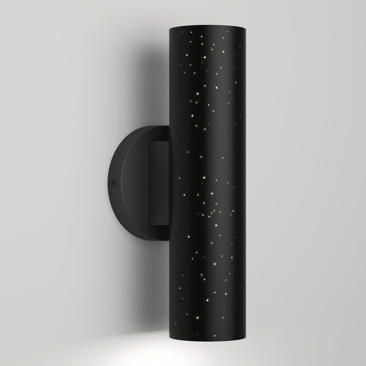 A modern black cylindrical wall sconce with small star-like holes, mounted on a white wall, emitting light from the top and bottom.