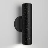 A modern black cylindrical wall sconce with small star-like holes, mounted on a white wall, emitting light from the top and bottom.