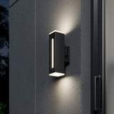 A modern, rectangular black outdoor wall light fixture emits a soft glow from both the top and bottom, mounted on a smooth gray wall near a glass door at night.