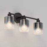A modern wall-mounted light fixture with three exposed bulbs, each covered by a cylindrical clear glass shade, set on a horizontal black metal bar against a white background.