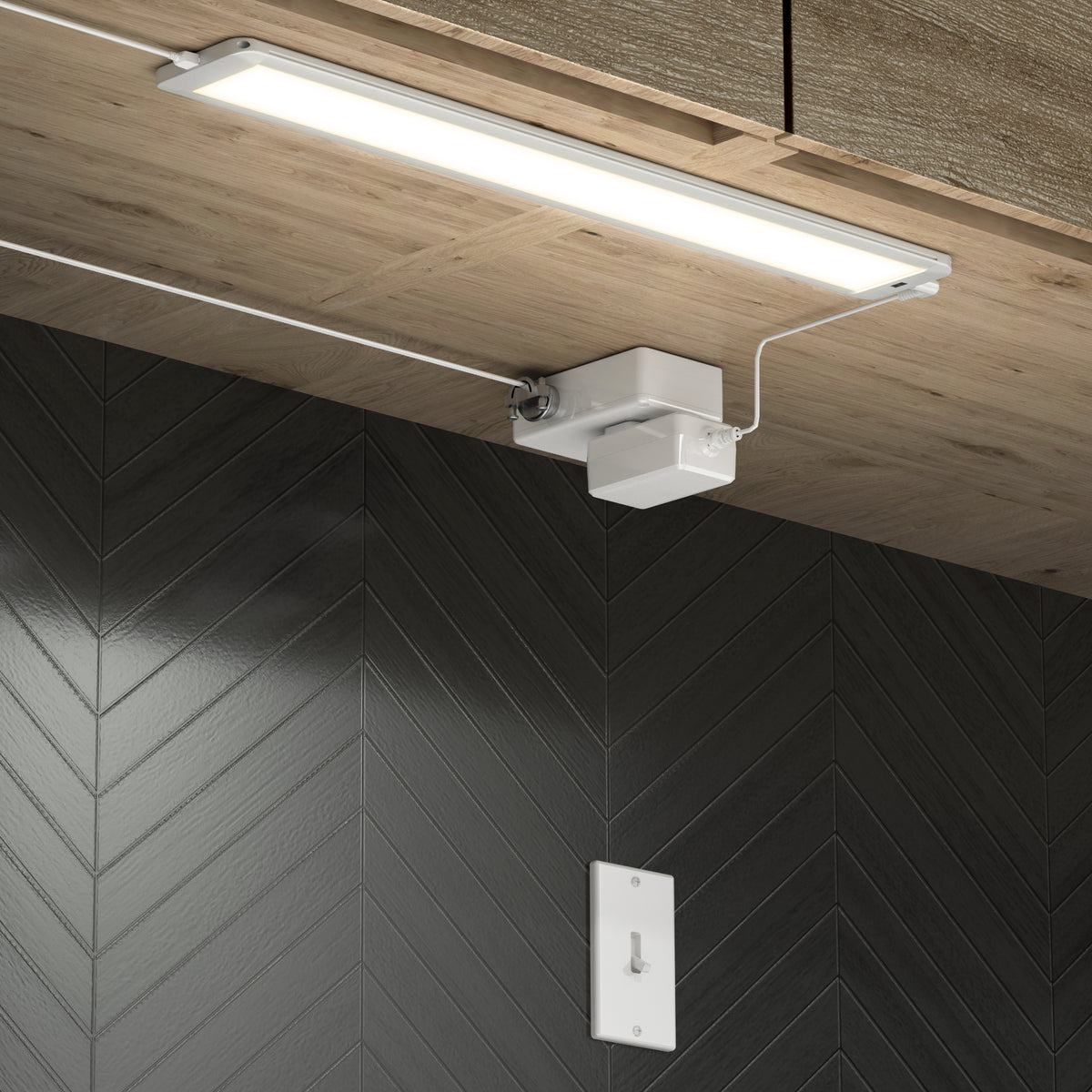A slim, rectangular LED light fixture is mounted under a wooden cabinet, illuminating a dark herringbone-patterned tile wall. Below the light, a white switch is installed on the wall.