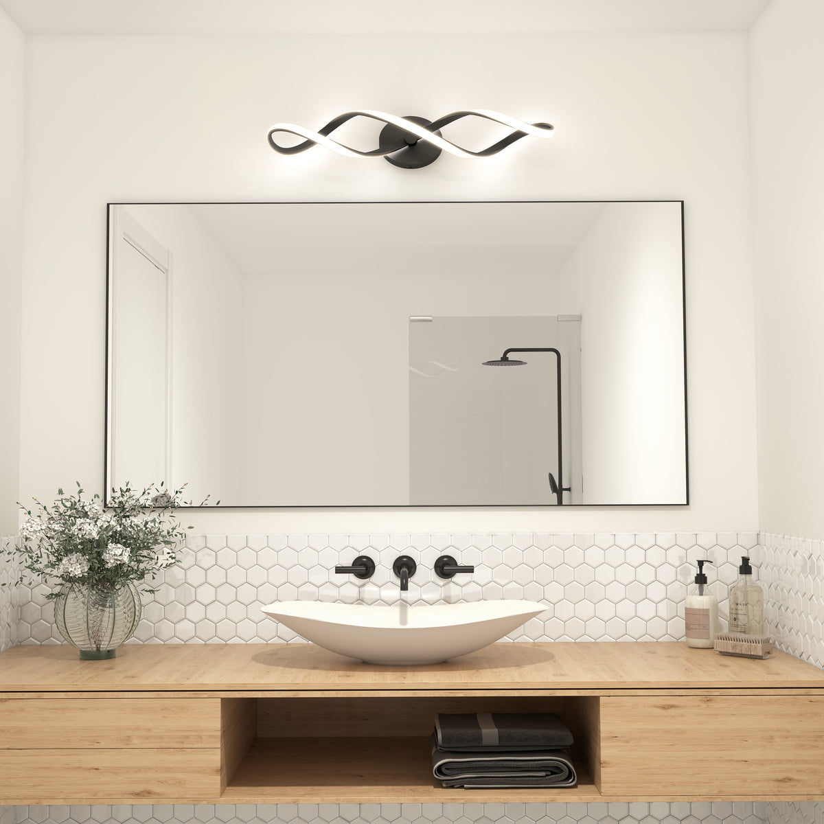 Modern bathroom featuring a large mirror, vessel sink, black fixtures, wood vanity, hexagon tile backsplash, vase with greenery, and the Artika Swirl LED bathroom vanity light above the mirror. Folded towels and toiletries sit on the counter.