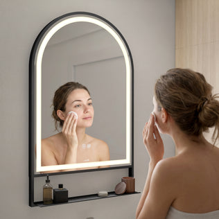 A woman wrapped in a towel applies a cotton pad to her cheek while looking at herself in the Artika Arturo 20 in. x 32 in. LED mirror with defogger, which is mounted above a shelf holding skincare products.