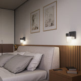 A modern bedroom with a cushioned bed, gray and white bedding, wooden headboard, two lit wall-mounted lamps, framed art above the bed, and a nightstand with books, a phone, and glasses.