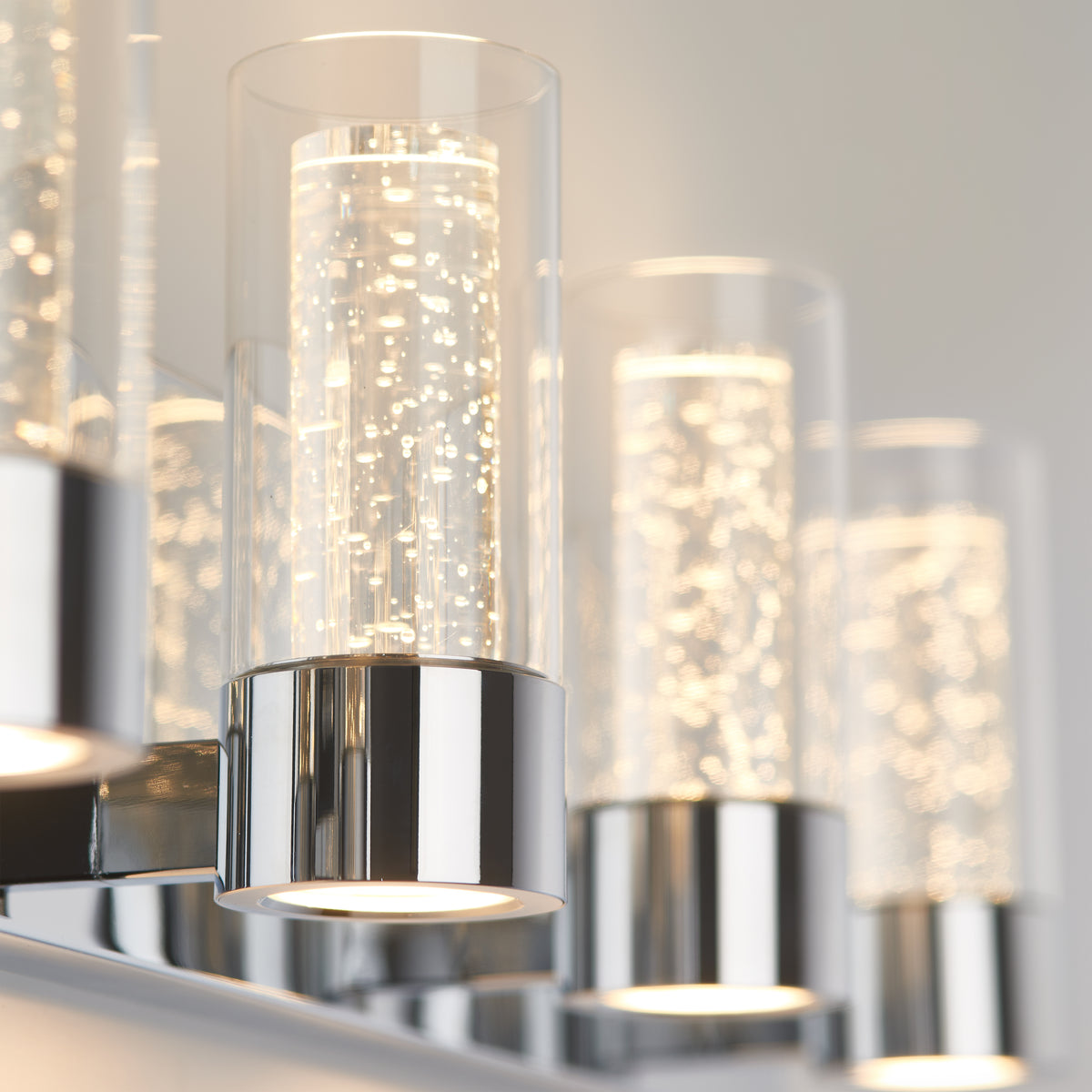 Close-up of modern cylindrical wall lights with chrome bases and clear glass shades containing bubbles, softly glowing against a neutral wall.