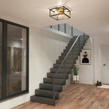 A modern interior features a black metal staircase with cable railing, a geometric ceiling light, wood flooring, a potted plant, abstract wall art, a large window, and a white door.