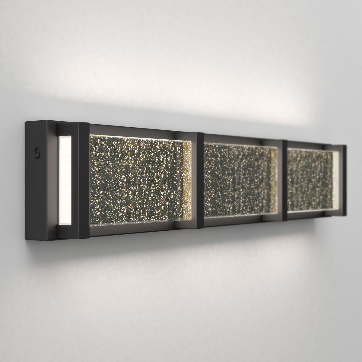 A modern wall-mounted light fixture with a rectangular black frame and three glass panels featuring a sparkling, textured surface, casting a soft glow onto a light-colored wall.