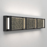 A modern wall-mounted light fixture with a rectangular black frame and three glass panels featuring a sparkling, textured surface, casting a soft glow onto a light-colored wall.