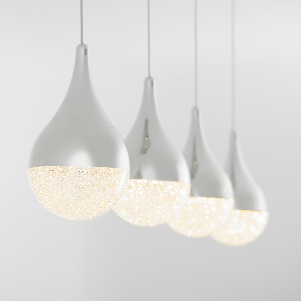 Four modern pendant lights with teardrop-shaped white fixtures hang in a row, glowing softly against a plain light grey background. The lower halves of the lights are textured and emit a warm, sparkling light.