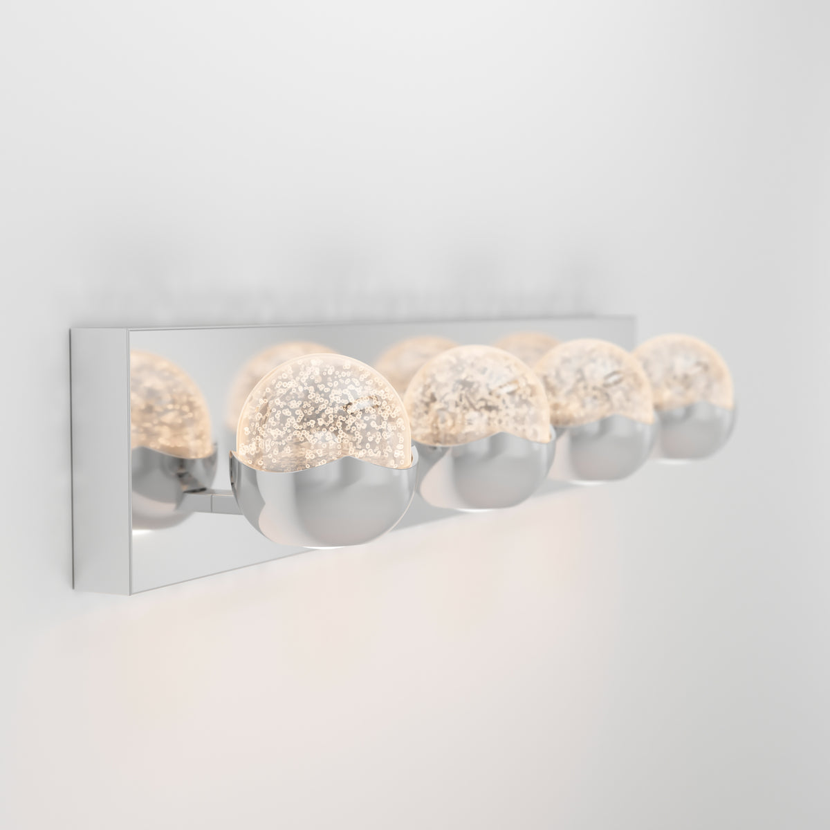 A modern wall-mounted light fixture with four round, half-chrome, half-textured glass bulbs attached to a shiny rectangular metal base, glowing softly against a white wall.