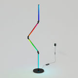 A modern, zigzag-shaped LED floor lamp with multicolor lights stands on a round black base, plugged into a wall outlet on a plain light gray background.