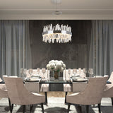 A modern dining room with a glass table set for six, beige upholstered chairs, a white floral centerpiece, and a contemporary chandelier hanging above. The backdrop features dark stone tiles and sheer curtains.