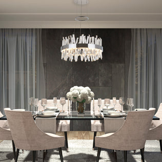 A modern dining room features a glass table for six, beige upholstered chairs, a white flower centerpiece, Artikas Enchanted LED pendant light, and floor-to-ceiling curtains against dark stone walls.