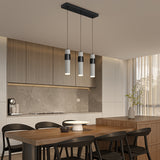 Modern kitchen with wooden dining table, black chairs, and three hanging pendant lights. Cabinets and backsplash are light wood and gray stone. Sunlight streams through sheer curtains by a vase with branches on the island.